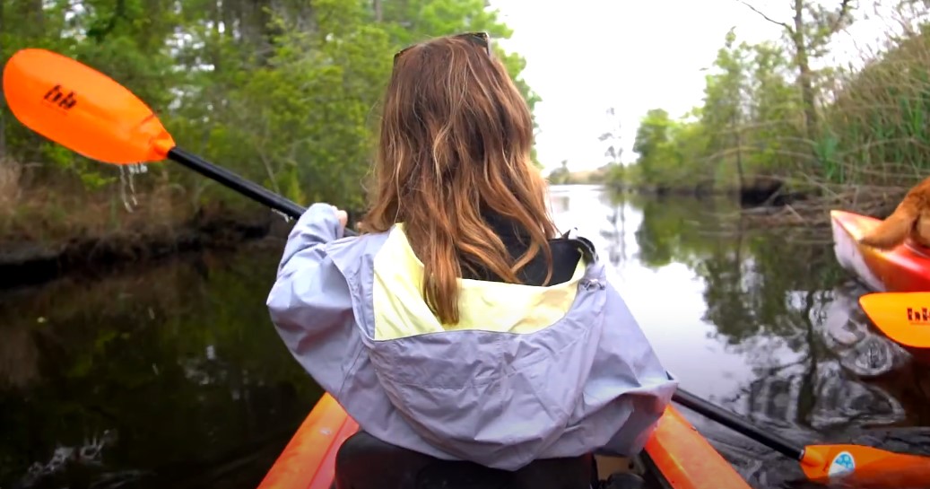 A woman paddles a kayak down a flowing river surrounded by lush greenery and sunlight