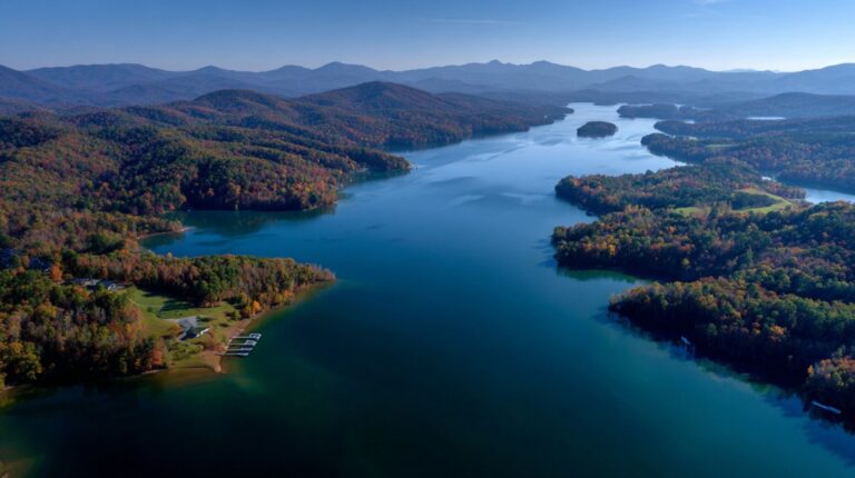 Aerial view of a long, winding lake surrounded by dense, colorful forest and distant blue mountains under a clear sky