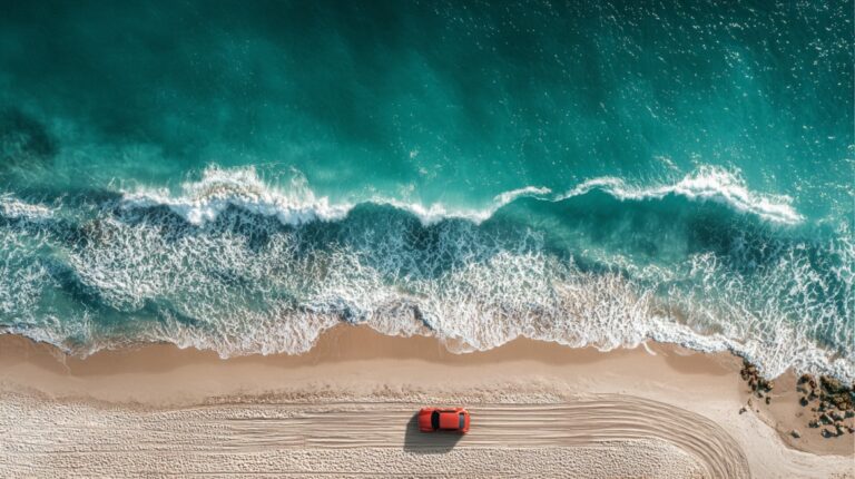 Aerial view of a red car parked on a sandy beach beside ocean waves