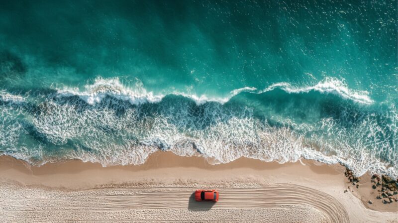 Aerial view of a red car parked on a sandy beach beside ocean waves